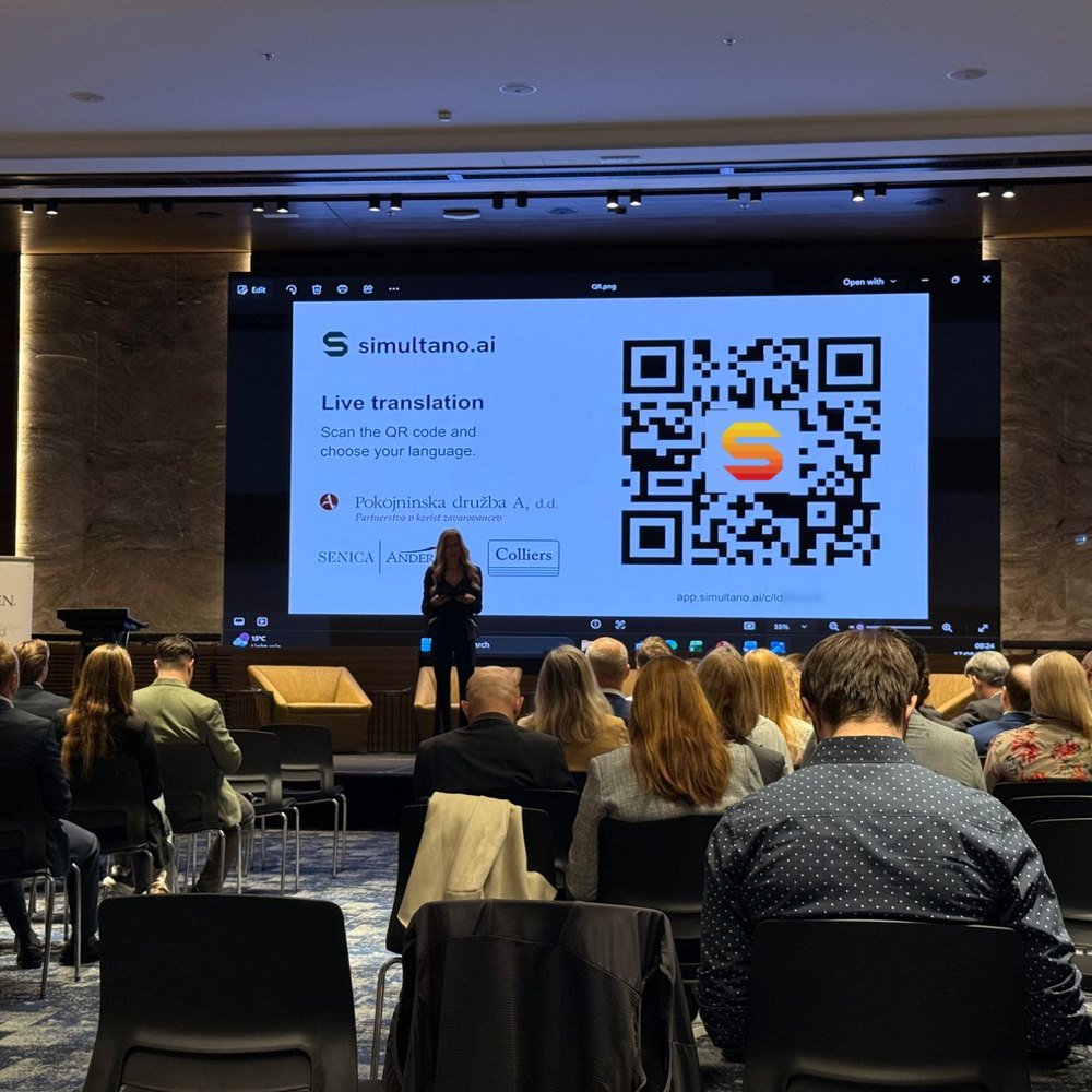 Konferencija u punoj sali: na platnu je simultano.ai s QR kodom, publika prati prevod na telefonima